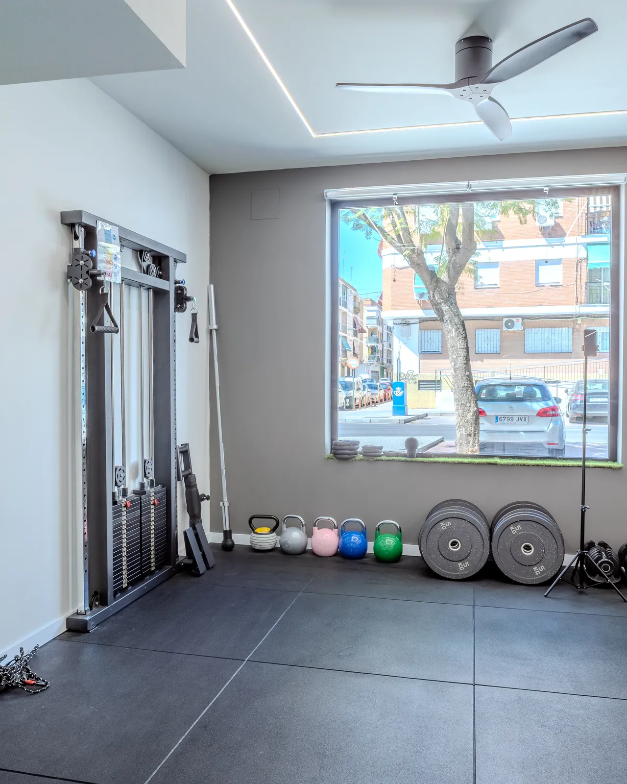 Sala de entrenamiento con ventanal a la calle, máquina de poleas, kettlebells y discos